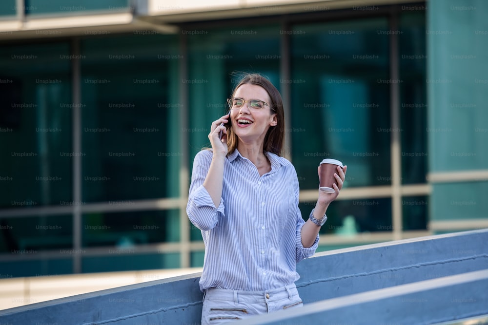 Attractive businesswoman drinking a coffee and using a mobile phone while standing at a window in an office overlooking the city