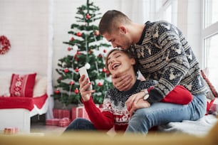 Guy kisses his girl in the head while she holds smartphone. Happy young people sits on the windowsill in the room with christmas decorations.