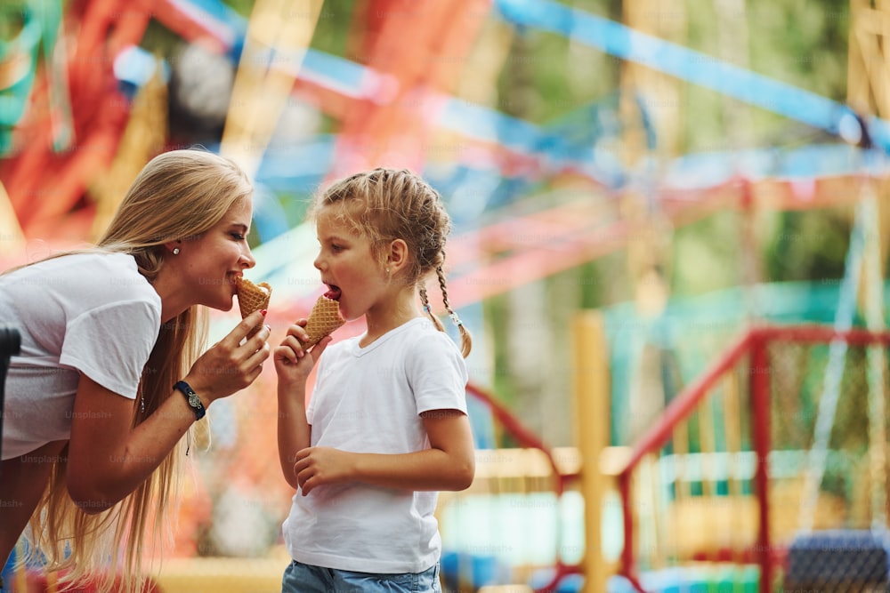 Comer helado. La niña alegre y su madre pasan un buen rato juntas en el parque cerca de las atracciones.