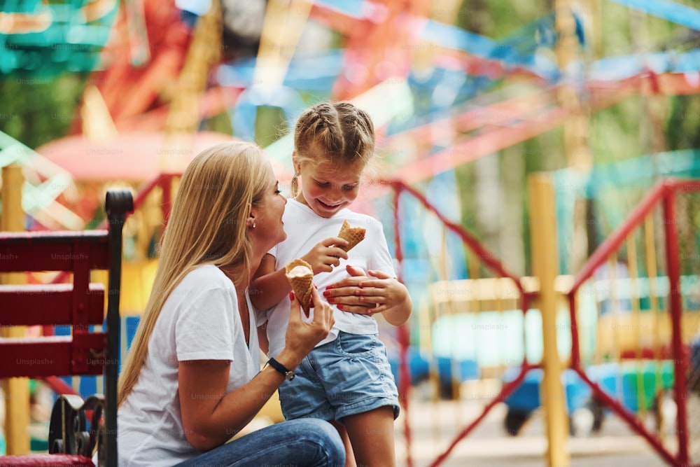 Comer sorvete. Menina alegre sua mãe se diverte no parque juntos perto de atrações.