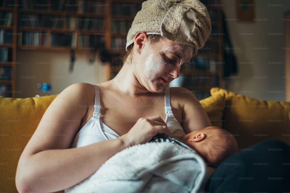 自宅で生まれたばかりの男の子に母乳を与え、フェイスマスクとタオルを頭にかぶった母親のショット