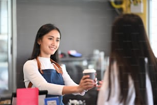 Young female waitress giving takeaway coffee cup to customers in coffee shop.