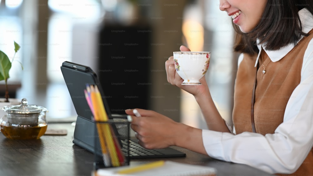 Abgeschnittene Aufnahme einer lächelnden Frau, die eine Kaffeetasse hält und mit einem Computer-Tablet arbeitet.