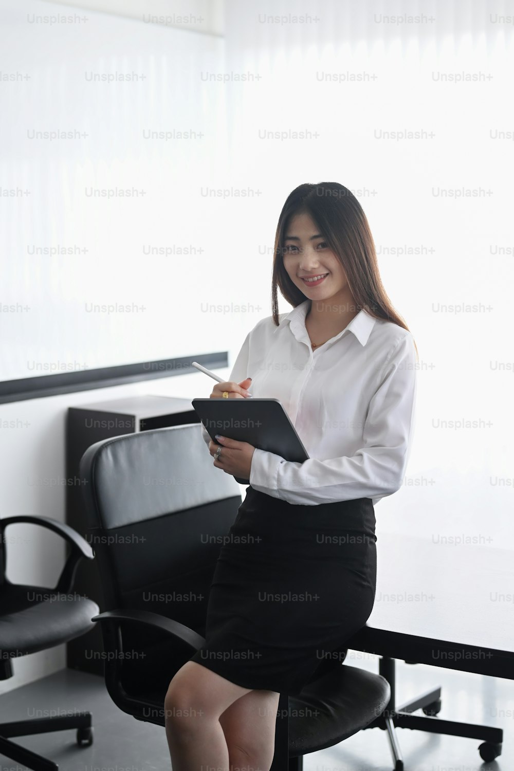 Portrait of young female office worker sitting on office desk and holding digital tablet.