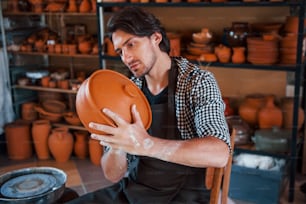 Young ceramist holds fresh handmade product made of clay in hands and looks at results of his work.