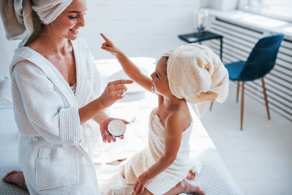 Usare la crema per schiarire la pelle. La giovane madre con la figlia ha una giornata di bellezza al chiuso in una stanza bianca.