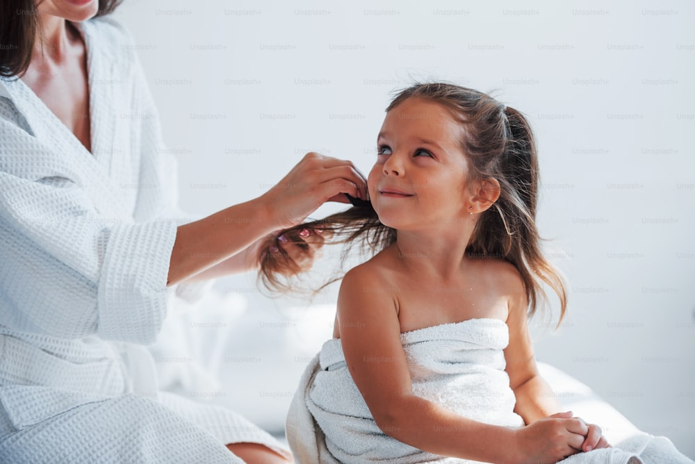 Aide à la coiffure. Une jeune mère avec sa fille a une journée de beauté à l’intérieur dans une salle blanche.