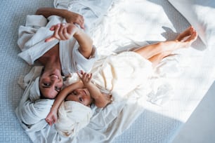 With hands up. Lying down on white bed. Young mother with her daugher have beauty day indoors in white room.
