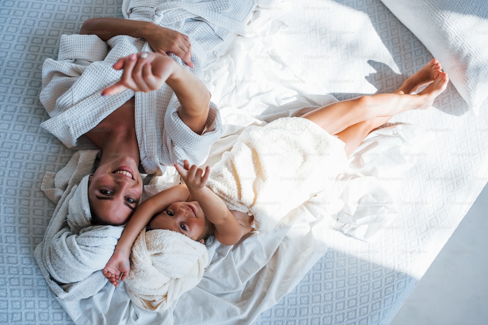 With hands up. Lying down on white bed. Young mother with her daugher have beauty day indoors in white room.