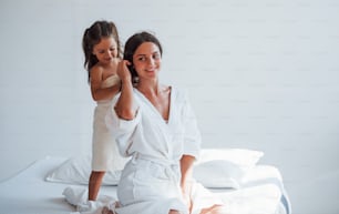 Helping to made hairstyle. Young mother with her daugher have beauty day indoors in white room.