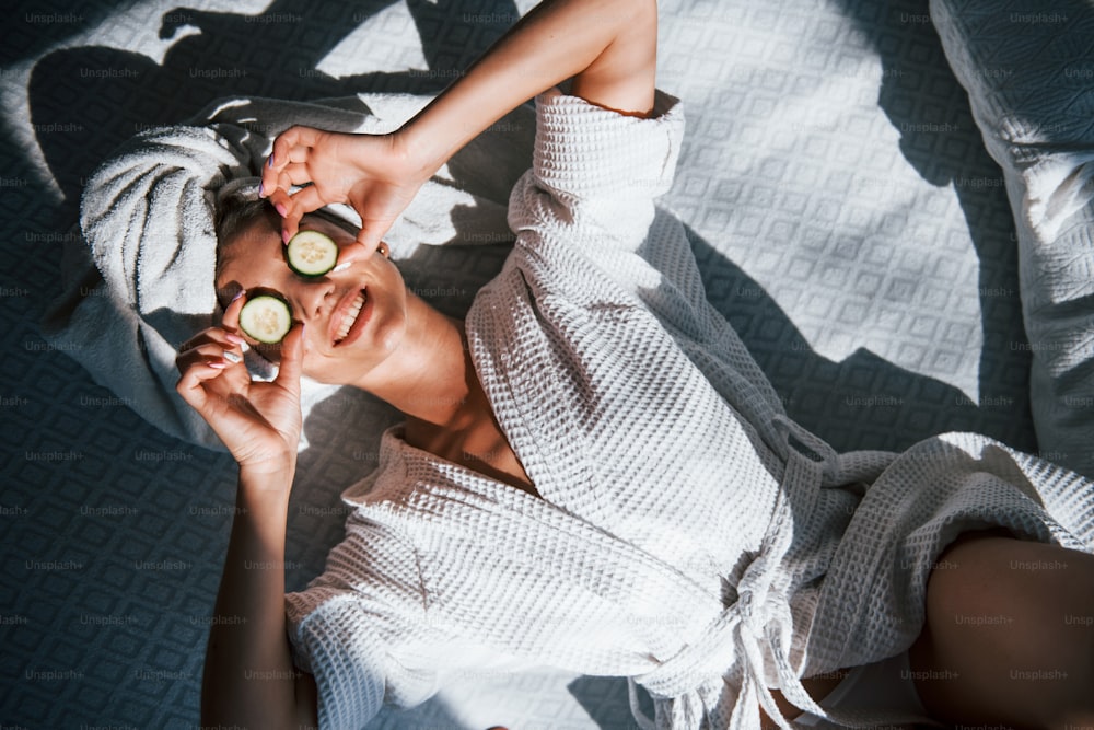 Cheerful mood. Young positive woman with towel on head lying on the bed with cucumber.