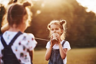 Two female kids stands in the field and talking by using string can phone.