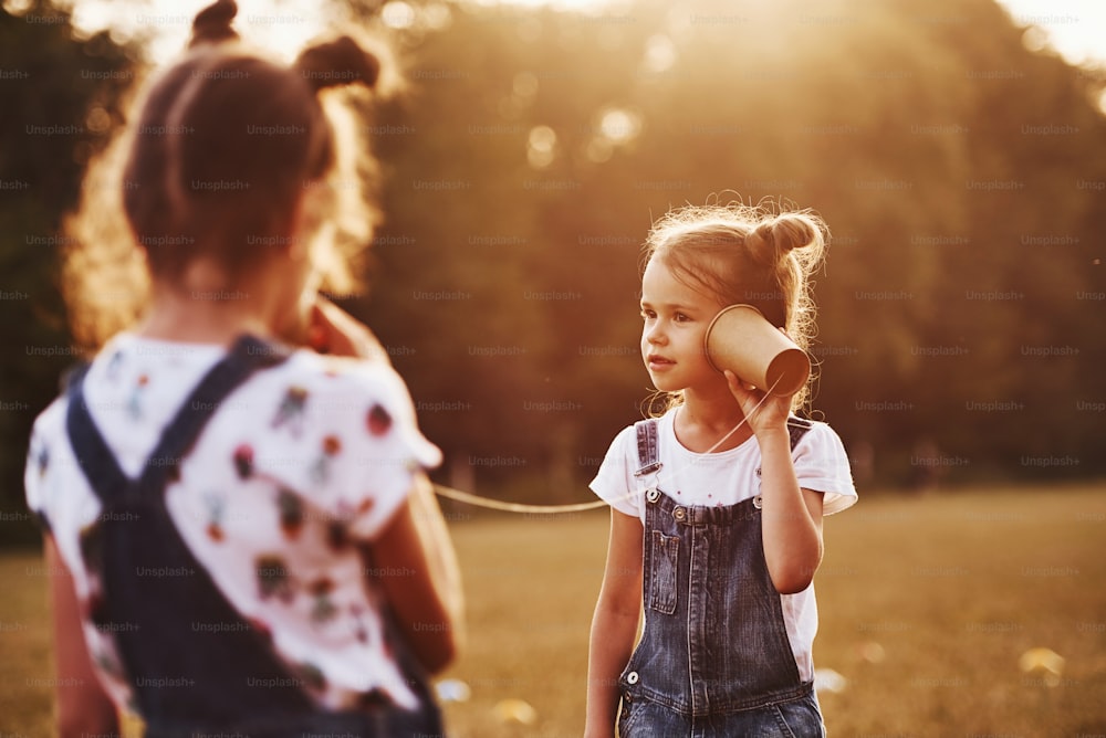 Two female kids stands in the field and talking by using string can phone.