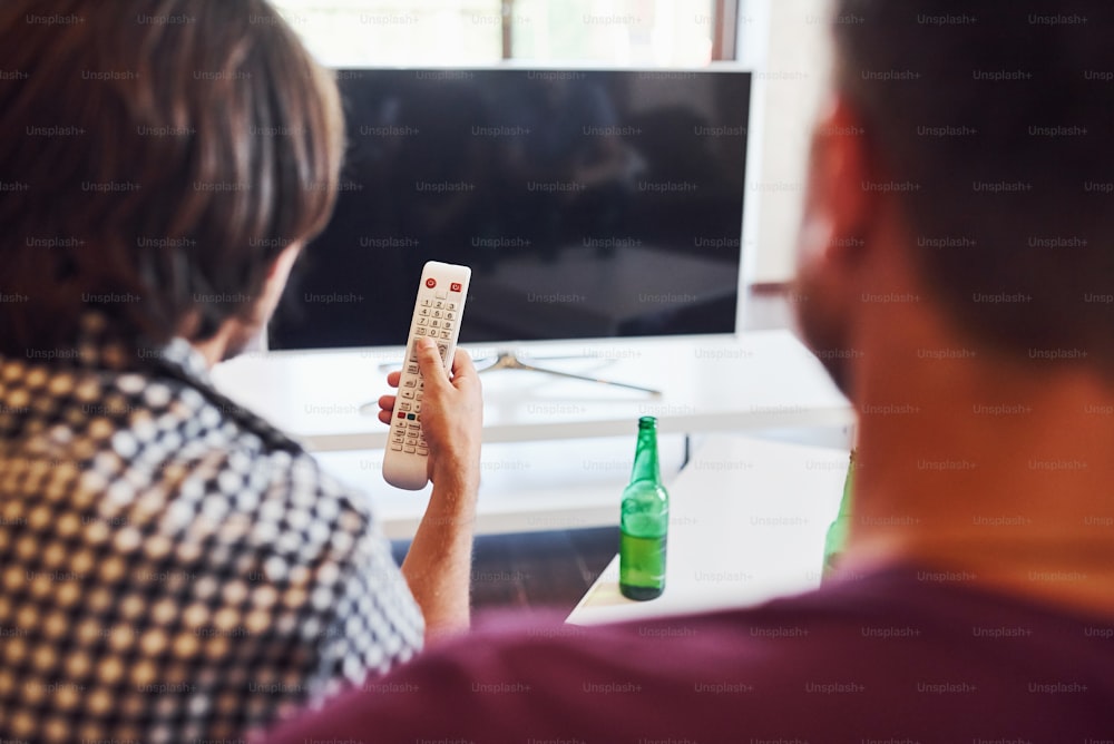 Rear view of friends watching TV and holds remote controller in hand.