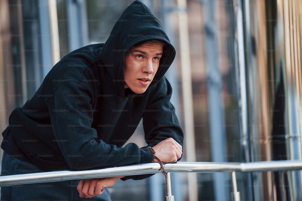 With hood on head. Portrait of young main in black clothes that leaning on the railings and taking a break.