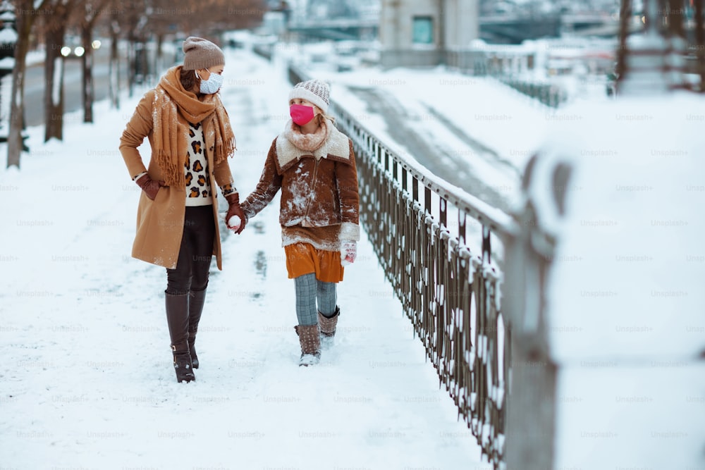 Full length portrait of stylish mother and daughter in a knitted hats and coats with medical mask walking outside in the city in winter.