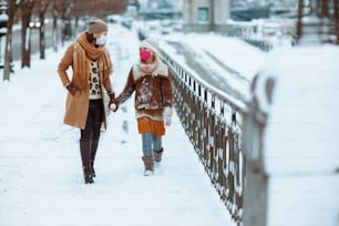 ニット帽とコートを着て、医療用マスクを着けたスタイリッシュな母と娘の全身肖像画が、冬の街を歩いている。