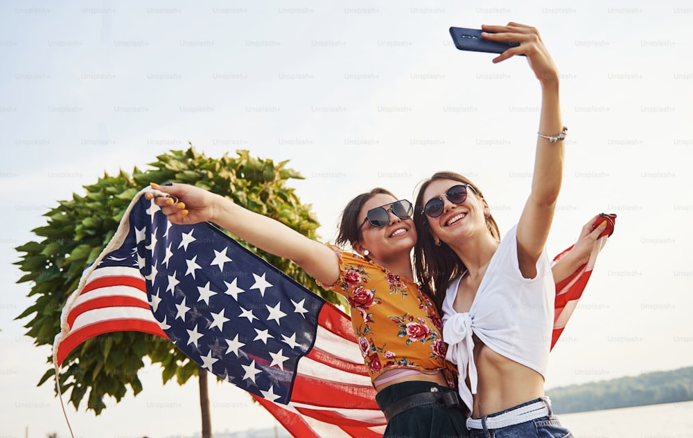 Green tree at background. Two patriotic cheerful women with USA flag in hands making selfie outdoors in park.