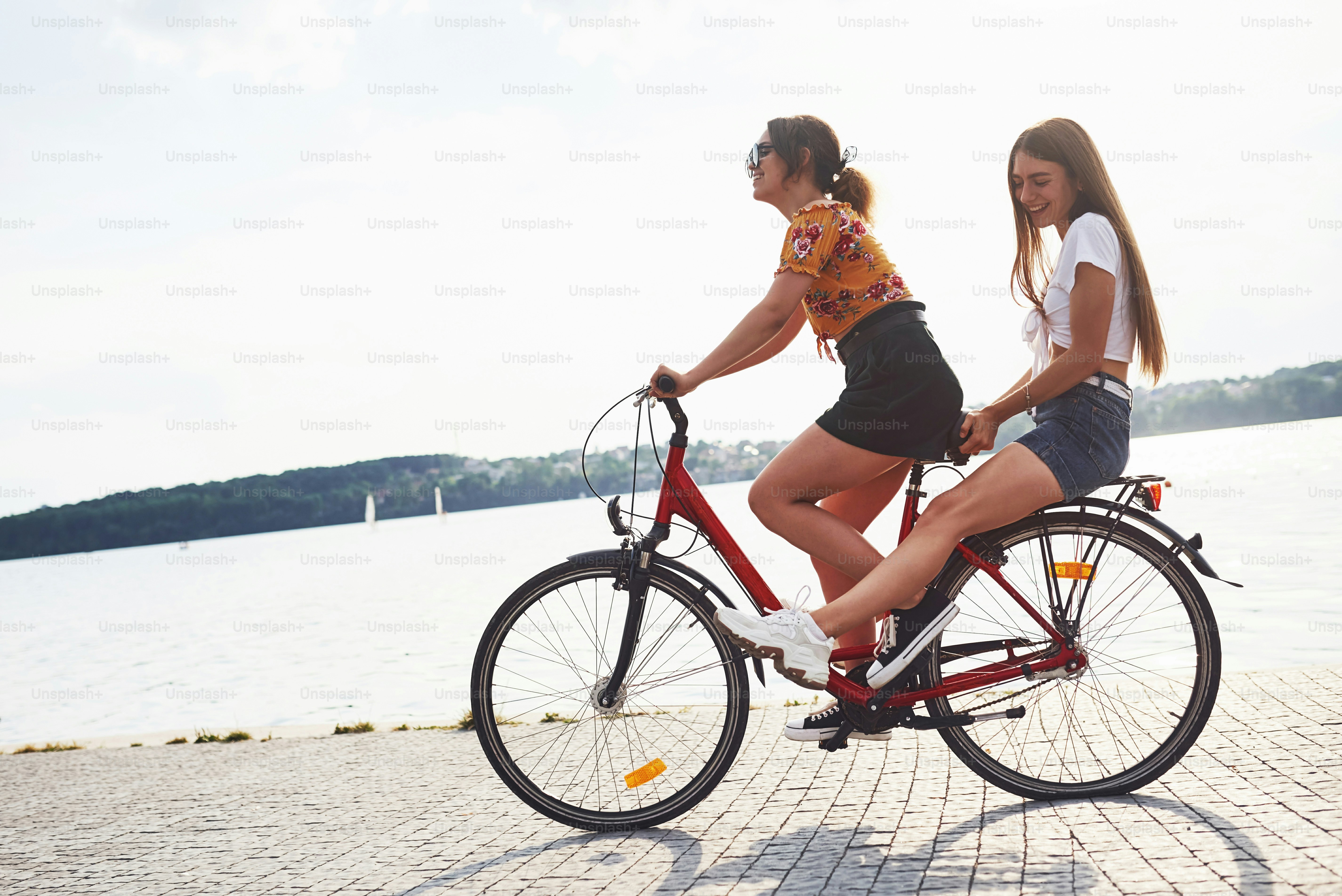 Two female friends on the bike have fun at beach near the lake.