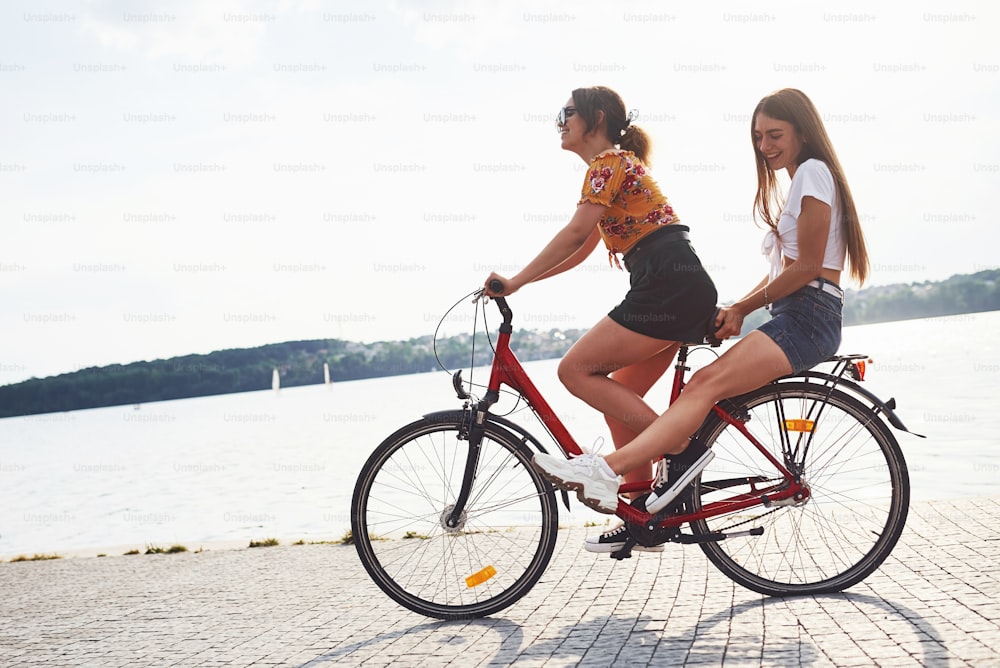 Two female friends on the bike have fun at beach near the lake.