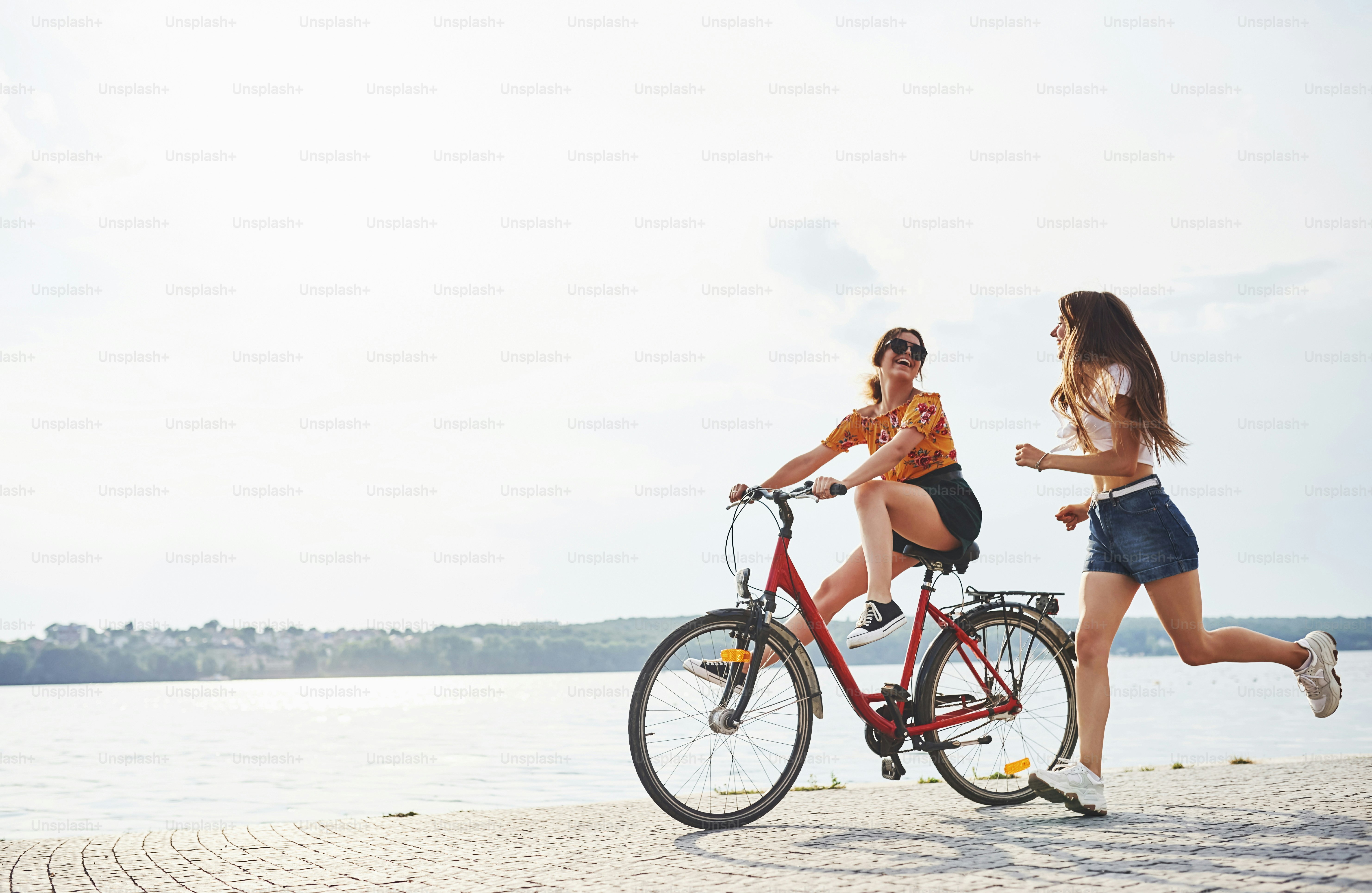 Girl runs near bicycle. Two female friends on the bike have fun at beach near the lake.