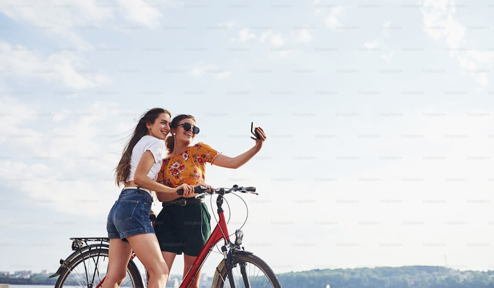 Tirando uma selfie incrível. Duas amigas na bicicleta se divertem na praia perto do lago.