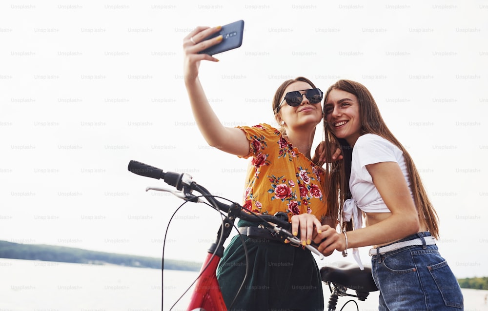 Making a selfie. Two female friends on the bike have fun at beach near the lake.
