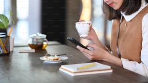 Cropped shot of woman holding a cup of tea and using mobile phone while sitting in cafe.