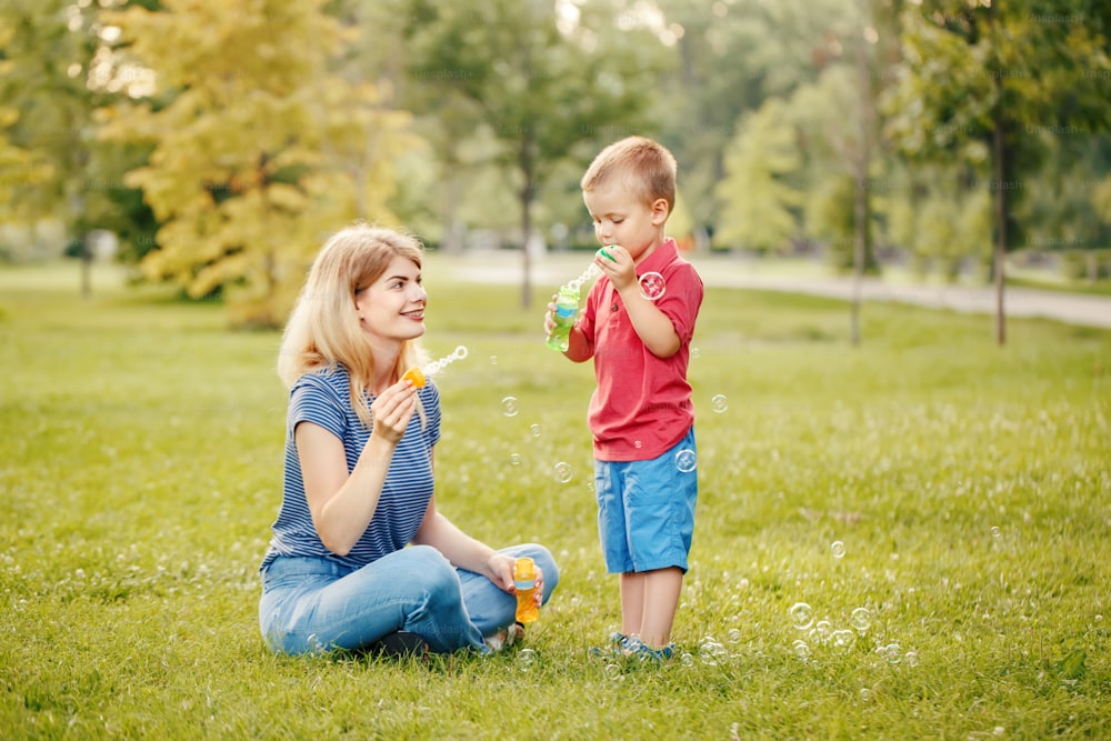 Young Caucasian mother and boy toddler son blowing soap bubbles in park. Mom and child playing having fun together outdoors on summer day. Happy authentic family childhood lifestyle.