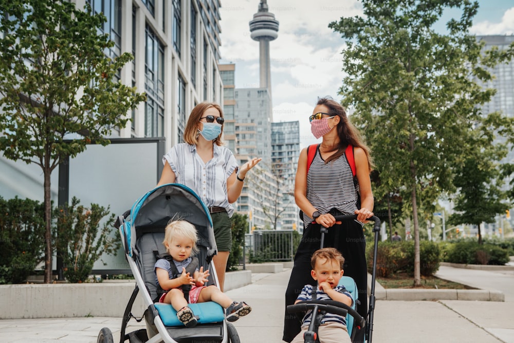 Two mothers walking down the street with kids in strollers. Friends in face masks chatting talking. People keeping the social physical distance outdoor during covid-19 coronavirus. New normal.