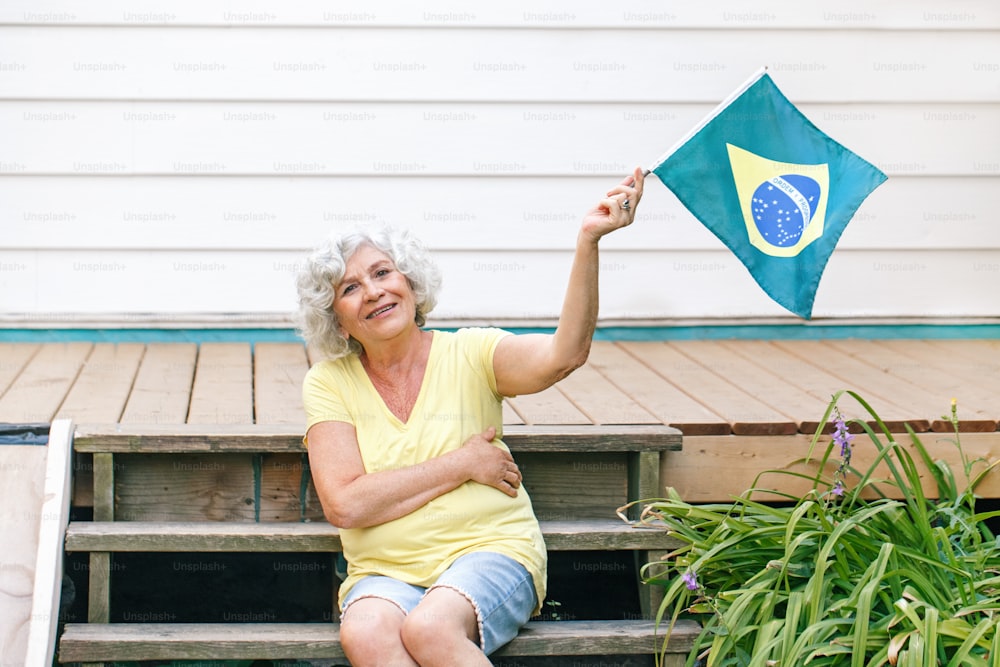 Stolzer Bürger, der den Unabhängigkeitstag Brasiliens feiert. Glückliche alte Frau, die die brasilianische Flagge im Freien hält. Lächelnde brasilianische ältere Dame, die auf dem heimischen Hinterhof sitzt und die brasilianische Flagge schwenkt.