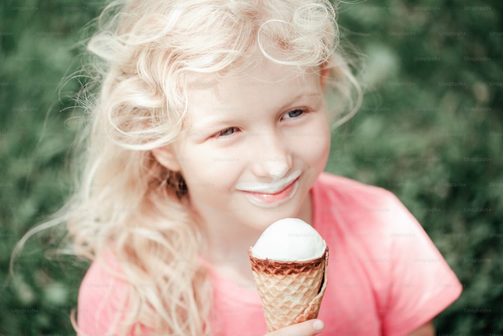 Cute funny adorable girl with long messy blonde hair sitting in park eating licking ice cream from waffle cone. Child eating tasty sweet cold summer food outdoors. Summer frozen meal snack.