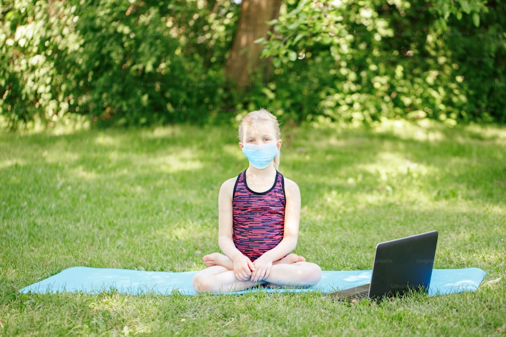 New normal. Yong girl child in face mask doing sport workout outdoors online. Video yoga class on Internet. Kid learning training in park with laptop. Social distance at coronavirus covid-19.