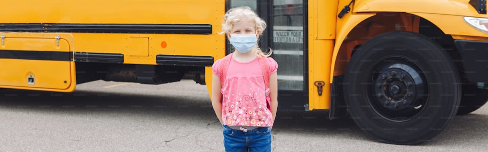 黄色いスクールバスのそばで青いフェイスマスクを着けた若い女の子の子供。教育と9月の学校への復帰。コロナウイルスcovid-19中のニューノーマル。Web バナー ヘッダー。