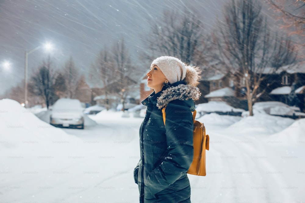 Woman in blue jacket winter clothes and hat walking outdoors under snow. Woman standing under street light looking at falling snow in the night evening. Winter romantic wonderland.