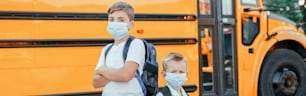 Brothers students kids wearing face mask near yellow bus. Education and back to school in September. New normal at coronavirus covid-19. Web banner header.