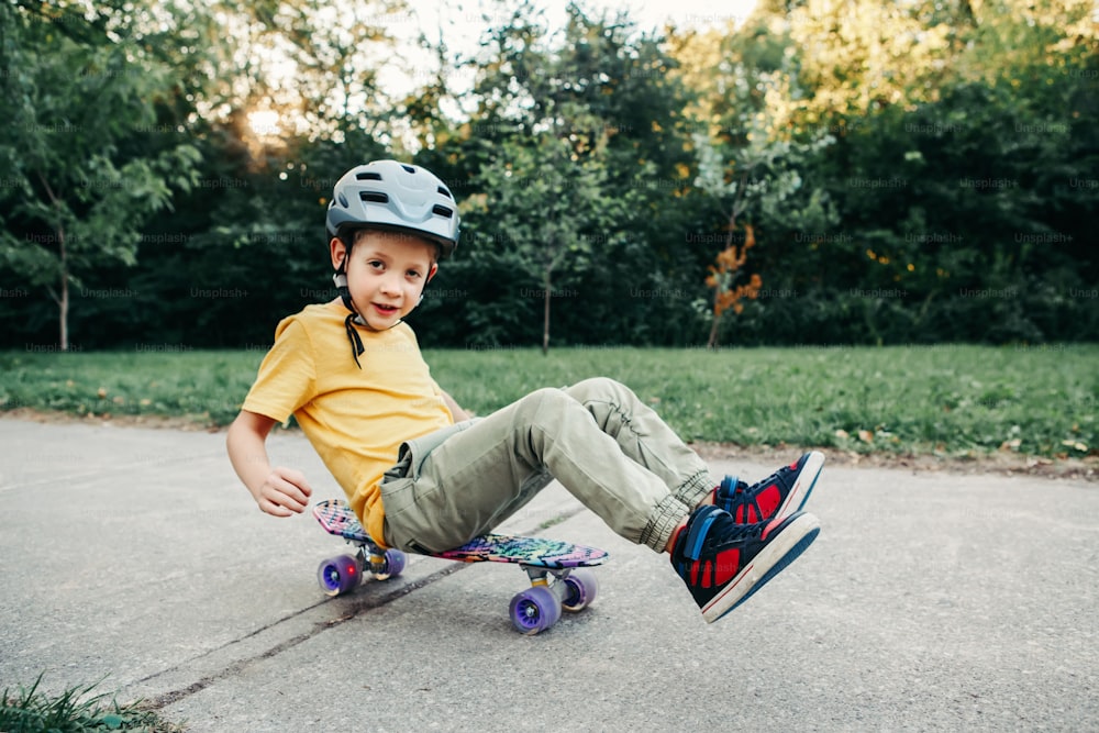 Happy Caucasian boy in grey helmet riding skateboard on road in park on summer day. Seasonal outdoor children activity sport. Healthy childhood lifestyle. Boy learning to ride skateboard.