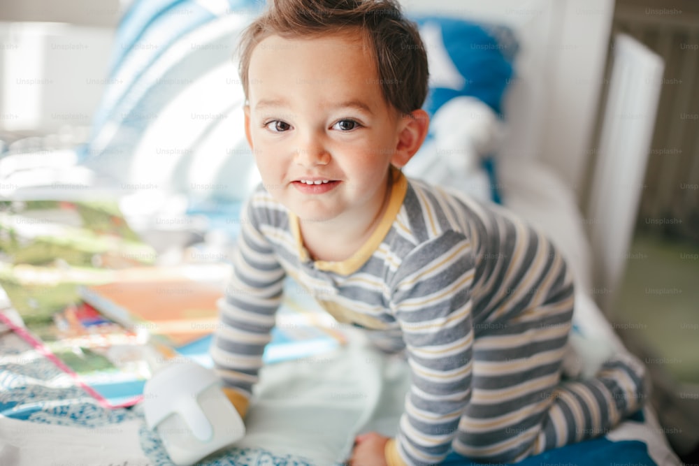 Cute adorable Caucasian kid boy sitting on bed drinking milk from kids bottle. Healthy eating drinking for little children. Supplementary food for growing babies. Candid real authentic moment.