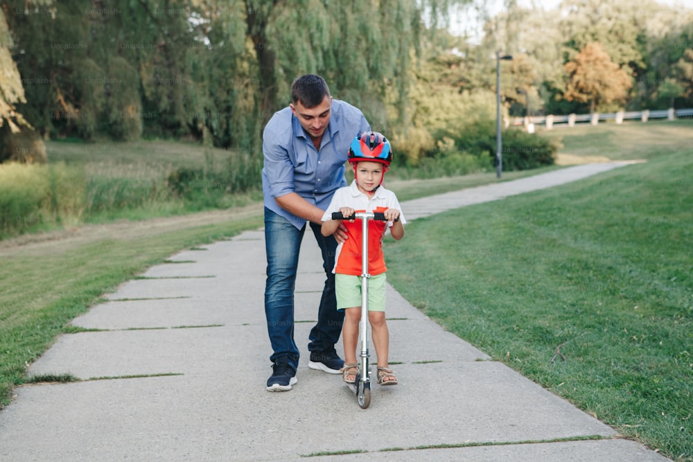 Père caucasien en train d’apprendre à son fils à faire du scooter. Enfant d’âge préscolaire en casque avec vélo sur la route du parc de l’arrière-cour. Activité saisonnière estivale pour les enfants en famille à l’extérieur.