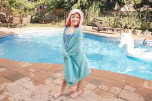 Cute adorable girl with freckles wrapped in beach towel standing by pool. Smiling funny child having fun in swimming pool. Summer outdoor water activity for kids.