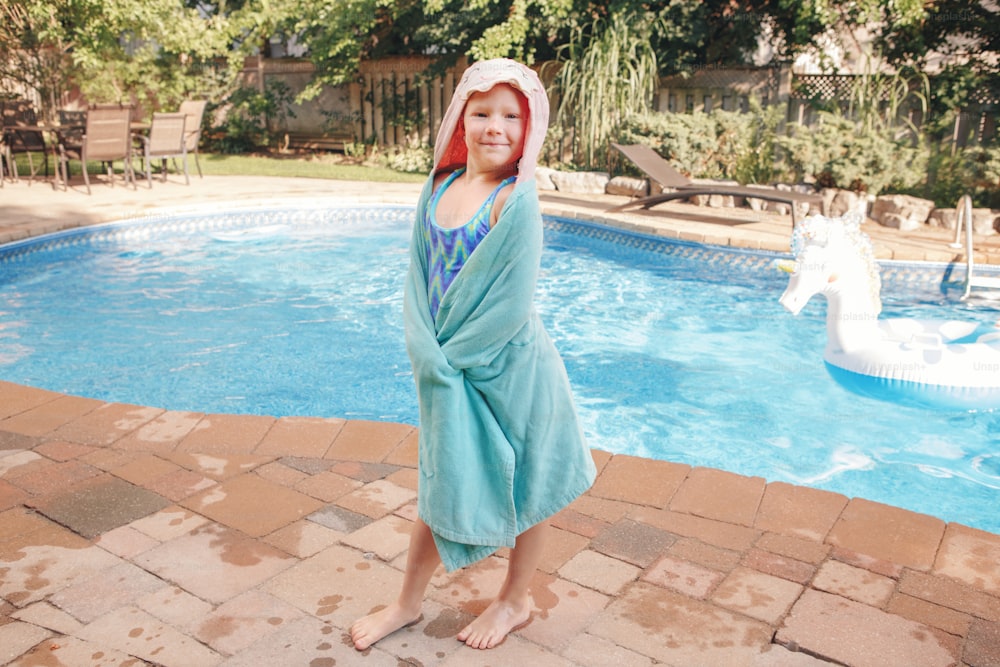 Cute adorable girl with freckles wrapped in beach towel standing by pool. Smiling funny child having fun in swimming pool. Summer outdoor water activity for kids.