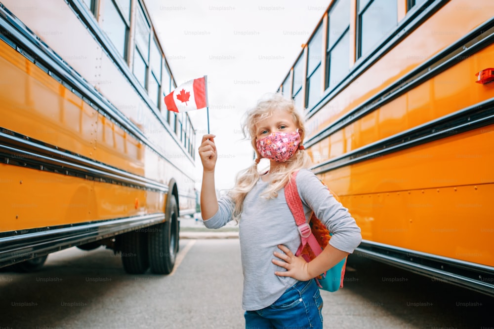 Caucasian girl student with face mask holding Canadian flag. Student kid near yellow school bus in Canada. Education and back to school in September. New normal during coronavirus.