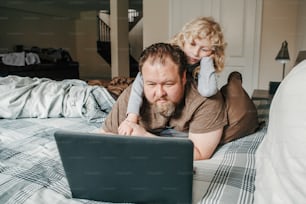 Work from home with kids children. Father working on laptop in bedroom with child daughter on his back. Funny candid family moment. New normal during coronavirus quarantine lockdown.