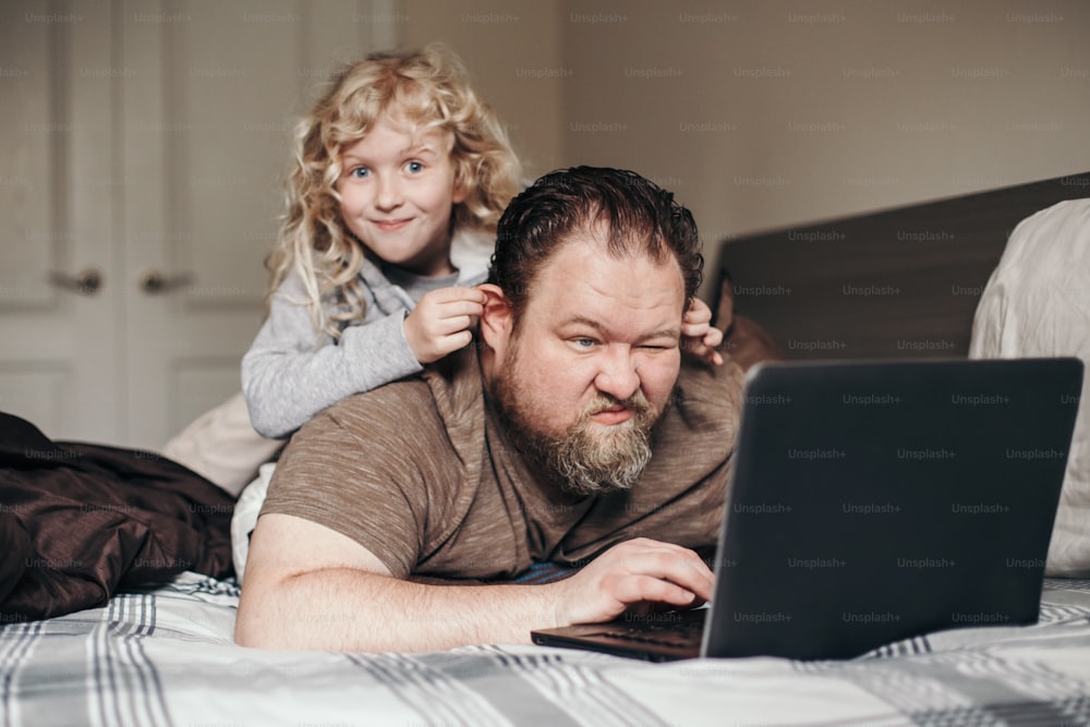 Work from home with kids children. Father working on laptop in bedroom with child daughter on his back. Funny candid family moment. New normal during coronavirus quarantine pandemic shutdown.