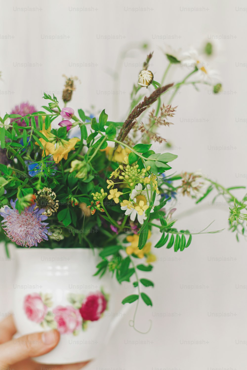 Foto Flores silvestres bonitas no copo vintage na mão da mulher no fundo do  pano rústico em casa. Olá Primavera. Feliz dia das mães e dia da mulher.  Colheita de flores –