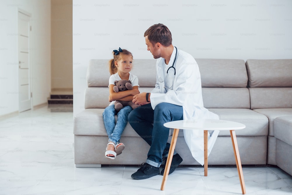 Männlicher Arzt in weißer Uniform sitzt in der Klinik mit kleinem Mädchen.