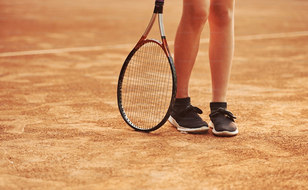 Legs and racket of female player that stands in the court at daytime.