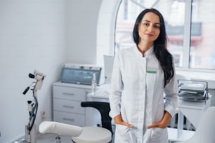 Brunette female doctor stands in modern office and looks at camera.