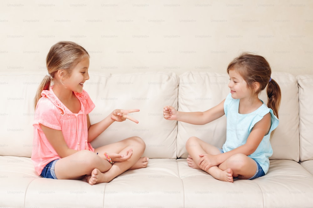 Duas amigas jogando jogo de mão de tesoura de papel de pedra. Crianças caucasianas sentadas no sofá brincando juntas. Interessante atividade de entretenimento para as crianças. Autêntico momento de estilo de vida cândido.