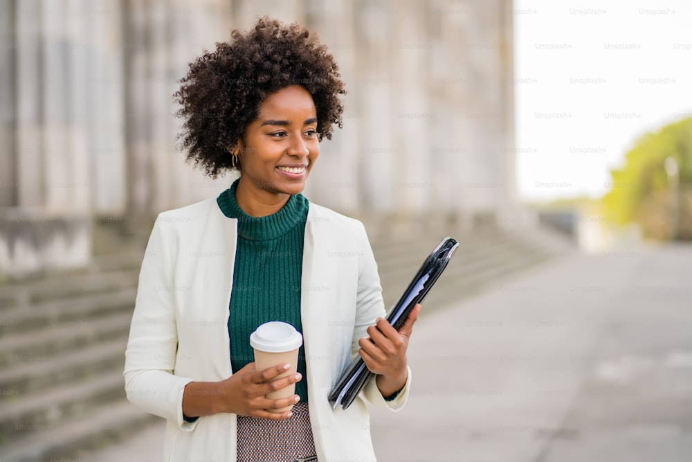 Portrait of afro businesswoman holding a cup of coffee and a clipboard while walking outdoors at the street. Business and urban concept.